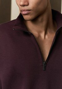 Burgunderfarbener Zip-up-Pullover mit hohem Kragen, ausgestattet mit einem schwarzen Reißverschluss und glatter Stoffoberfläche, zeigt eine Nahaufnahme des Halsauschnitts.