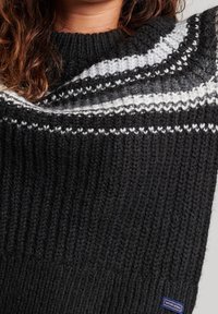 Schwarzer Strickpullover mit einem strukturierten Stoff und weißen sowie grauen kreisförmigen Mustern am Ausschnitt; mit Markenetikett unten links.