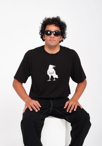 Zwarte T-shirt met een witte kraaienafbeelding en de tekst "VULCAN." Draagt het met zwarte broeken. Model zit op een witte kubus.