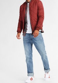 Prošívaná maroon bomber bunda, bílá tričko, kostkovaná košile pod ní, světle modré džíny a bílé tenisky s červenými pruhy.