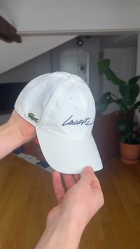 Käed hoiavad valget Lacoste'i pesapalli mütsi, millel on tikitud logo ja roheline krokodilli embleem, ning toas on puidust põrand ja potitaim.