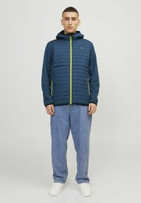 Giacca trapuntata navy con cappuccio, caratterizzata da una zip completa e dettagli verde brillante. Indossata sopra jeans baggy azzurri e sneaker bianche.