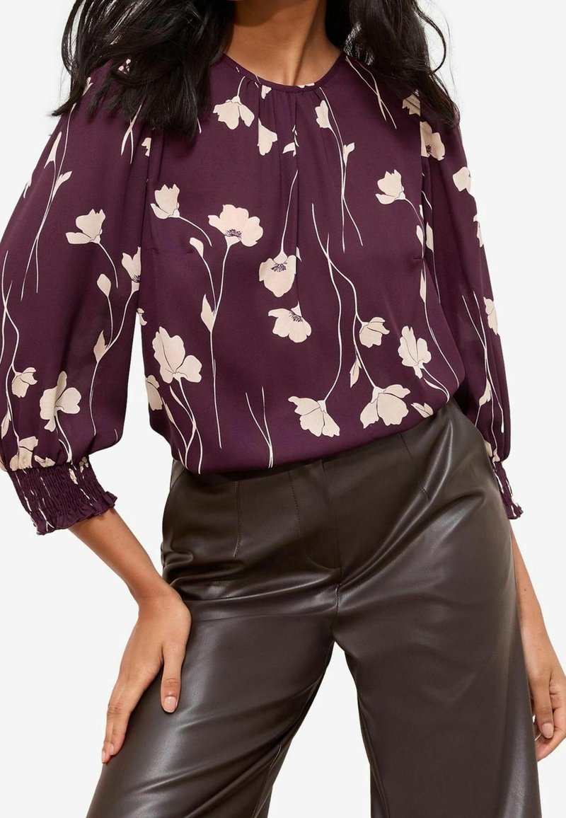 Femme portant un chemisier violet à motifs floraux avec des manches longues et un pantalon en cuir marron foncé, main posée sur la hanche.