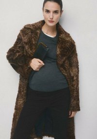 Kobieta w brązowym futrzanym płaszczu z sztucznego futra, nałożonym na szary sweter i czarne spodnie, trzymająca czarną kopertówkę na jednolitym tle.