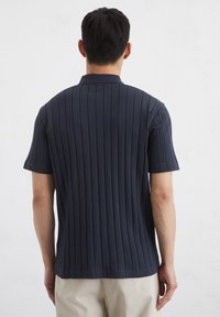 Marineblauw polo shirt met korte mouwen en verticale ribbelstructuur. Heeft een omslaande kraag en een rechte zoom. Achteraan zichtbaar met strakke lijnen en naden.