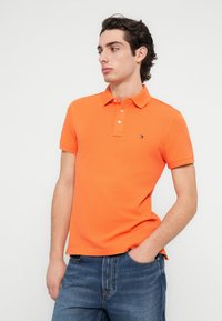 Πορτοκαλί πόλο μπλούζα με γιακά, με κουμπιά και ένα μικρό λογότυπο στο στήθος. Φορεμένη με μπλε τζιν παντελόνι.