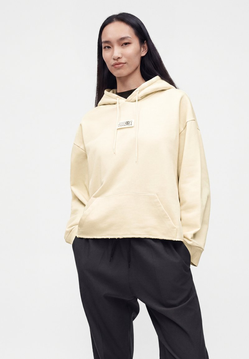 Femme aux longs cheveux noirs portant un sweat à capuche beige clair avec poche avant et un pantalon noir, debout devant un fond blanc uni.