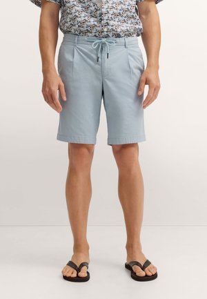 Mann trägt hellblaue Shorts mit Kordelzug, blumiges kurzärmliges Hemd und schwarze Flip-Flops, steht vor einem einfarbigen Hintergrund.