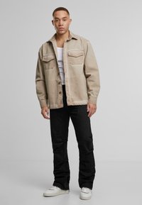 Muž stojaci v béžovom kbuttonovom saku, bielom tielku, čiernych nohaviciach a bielych teniskách na pozadí jednoduchého šedého pozadia.