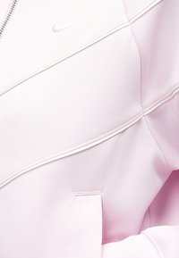 Ροζ ανοιχτό φούτερ Nike με διαγώνιες ραφές, μπροστινή τσέπη και μικρό λευκό λογότυπο Nike στο στήθος.