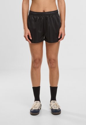 Persoon die zwarte sportshorts, zwarte kousen en blauw-witte Adidas-sneakers draagt, staand op een effen grijze achtergrond.
