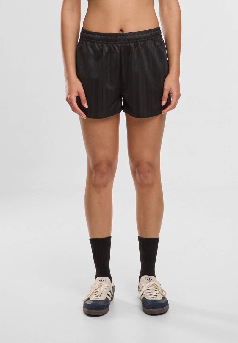 Personne portant un short de sport noir, des chaussettes montantes noires et des baskets Adidas bleues et blanches, debout sur un fond gris uni.
