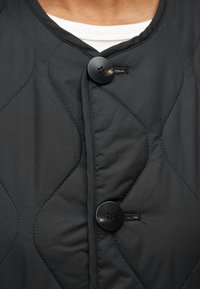 Μαύρο καπιτονέ μπουφάν με στρογγυλή λαιμόκοψη, δύο μαύρα κουμπιά και λεία υφή. Περιλαμβάνει ορατά μ οτίβα ραφής.