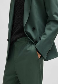 Veste de costume verte avec des boutons noirs et des revers crantés, associée à un pantalon assorti avec une taille élastique et un pull noir texturé en dessous.