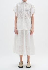 Completo in cotone bianco a due pezzi, composto da una camicia a maniche corte con bottoni e una gonna al ginocchio, entrambi con dettagli ad occhiello e una vestibilità morbida.