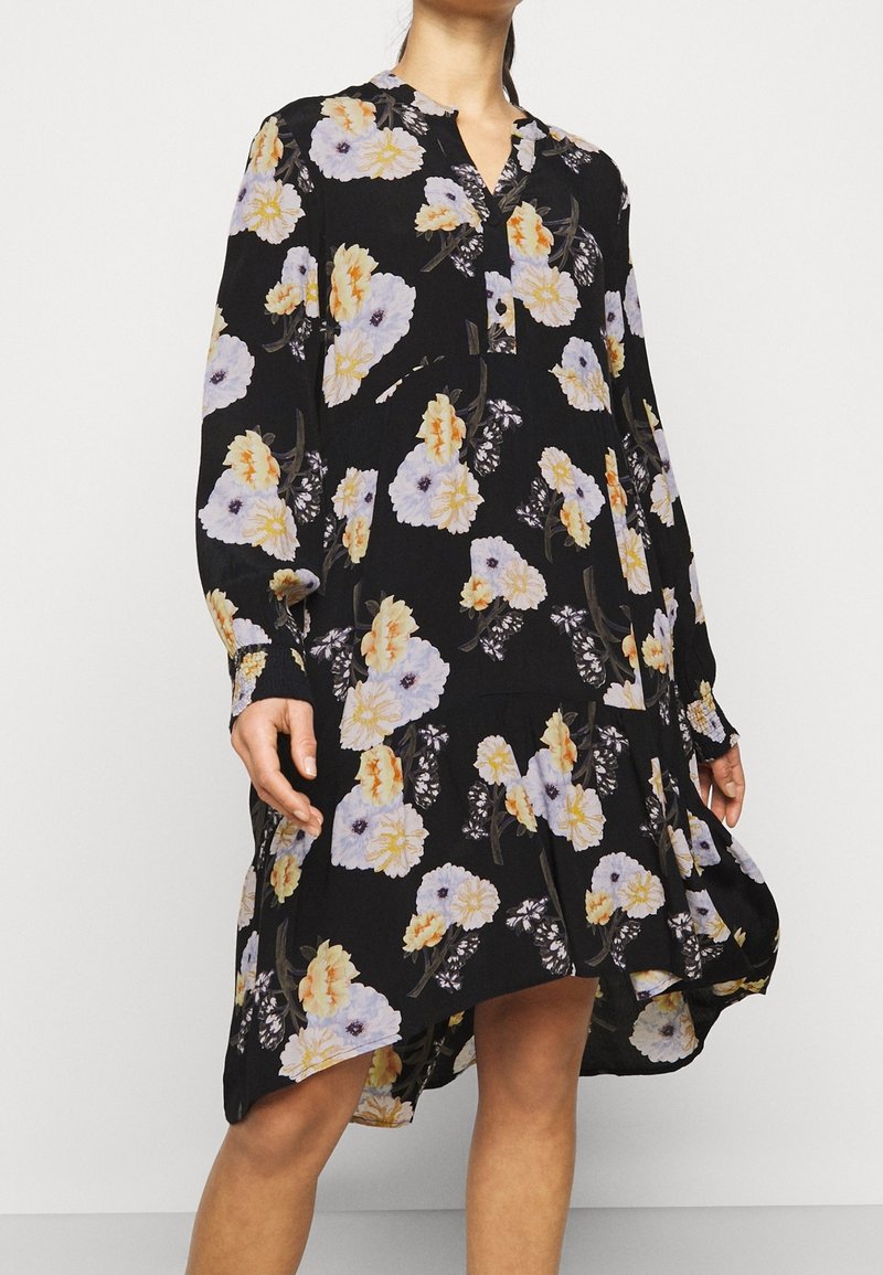 Černé květinové šaty s dlouhými rukávy, které zdobí směs bílých, žlutých a fialových květin. Lehce průsvitná tkanina s pohodlným střihem a zakřiveným spodním lemem.