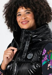 Schwarze Steppweste mit hohem Kragen, glänzender Oberfläche, Reißverschluss mit Markenband und Logo-Label auf der Vorderseite. Bunte Blumenakzente sind sichtbar.