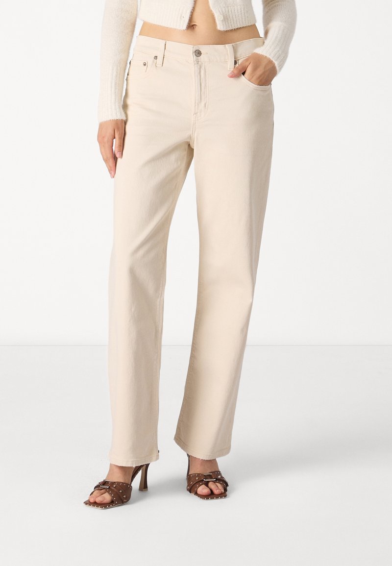 Pantaloni beige in cotone a gamba dritta con vita media, tasche anteriori e una leggera trama. Abbinati a sandali con tacco marroni.