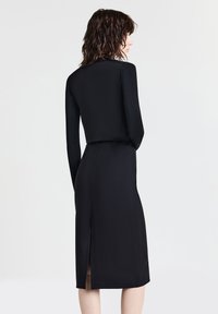 Donna con capelli scuri alla spalla, che indossa un top fitted a maniche lunghe nero e una gonna a matita abbinata con uno spacco sul retro, rivolta verso l'esterno.