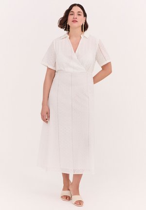 Femme aux cheveux courts et foncés portant une robe midi blanche à œillets avec des manches courtes et des sandales beiges à bout ouvert, debout devant un fond uni.