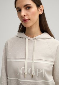 Lichtbeige hoodie met een zachte textuur, met een subtiel patroon en "JOOP!" geborduurd in een contrasterende kleur. Touwtjes met gouden accenten.