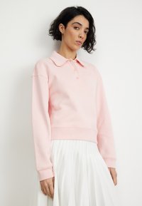 Sudadera de color rosa claro con cuello, tres botones y puños acanalados, llevada sobre una falda blanca con plisados. Textura suave y diseño informal.