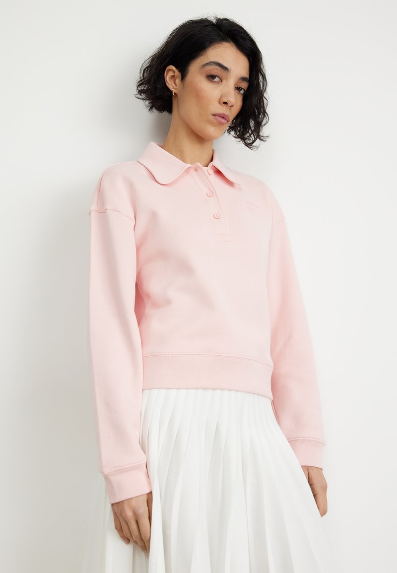 Sudadera de color rosa claro con cuello, tres botones y puños acanalados, llevada sobre una falda blanca con plisados. Textura suave y diseño informal.