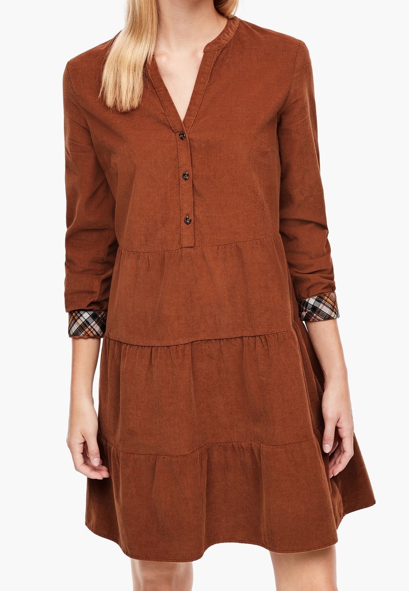 Femme portant une robe longue à manches longues marron à étages, avec un col en V, trois boutons et des poignets à motif à carreaux, debout devant un fond blanc.