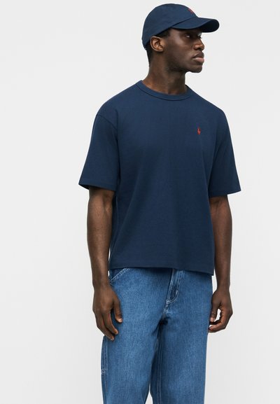 Άνδρας που φοράει μπλε κοντομάνικο μπλουζάκι, αντίστοιχο καπέλο και μπλε τζιν, στέκεται και κοιτάζει προς τα δεξιά μπροστά σε απλό φόντο.