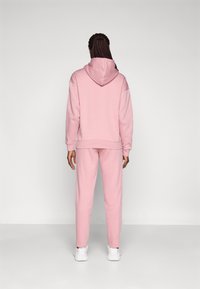 Roze sweatshirt met capuchon, losse pasvorm en ribgebreide manchetten; bijpassende roze joggingsbroek met zijpanelen. Draagt met witte sneakers.