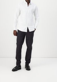 Camicia bianca a maniche lunghe con bottoni abbinata a pantaloni a quadretti scuri con linee rosse. Le scarpe sono mocassini neri.