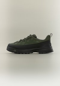 Scarpa da trekking verde scuro e nera con suola spessa, design con lacci e linguetta sul tallone, posizionata su uno sfondo neutro.