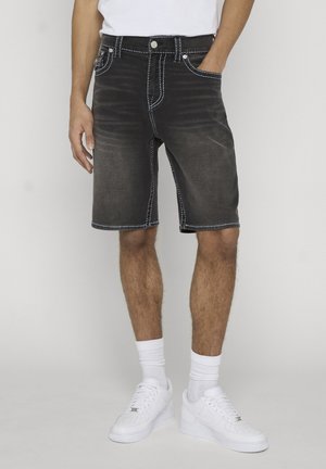 Man draagt vervaagde zwarte spijkershorts, witte sokken en witte sneakers, staat met één hand in de zak.