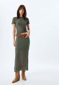 Vestido de punto acanalado en verde oliva con textura ondulada, mangas cortas y un cinturón marrón. Combinado con botas vaqueras en color beige.