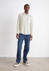 Camisa abotonada color beige claro con un solo bolsillo en el pecho, combinada con jeans rectos de color azul oscuro y zapatillas en blanco y negro.
