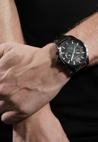 Juodas chronografas su tekstūruota juoda odinė apyranke, metaliniais akcentais, tamsiu ciferblatu ir trimis subdailiais; dėvimas ant riešo.