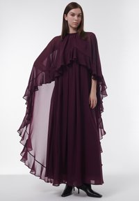 Burgunderfarbene Chiffonkleid mit schichten Rüschen, fließenden Kapuzenärmeln und hohem Kragen, kombiniert mit schwarzen High-Heel-Stiefeln.