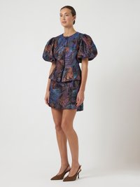 Donna che indossa un vestito metallico a fantasia con maniche ampie e un minidress, abbinato a tacchi slingback marroni, in piedi davanti a uno sfondo bianco.