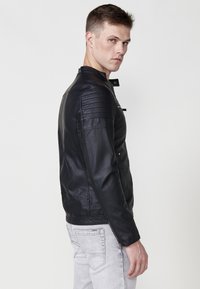 Chaqueta de piel sintética negra con cuello alto, detalles acolchados en los hombros y bolsillos con cremallera, combinada con pantalones de mezclilla gris claro.