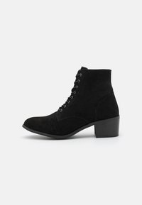 Botas de tornozelo em camurça preta com bico arredondado, atacadores na frente e um salto baixo quadrado. Apresentam uma textura suave e ilhós prateados.