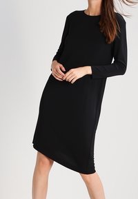 Nainen, jolla on polvimittainen musta pitkähihaisuus mekko, seisoo hieman kääntyneenä ja kädet ristissä edessään yksinkertaista taustaa vasten.