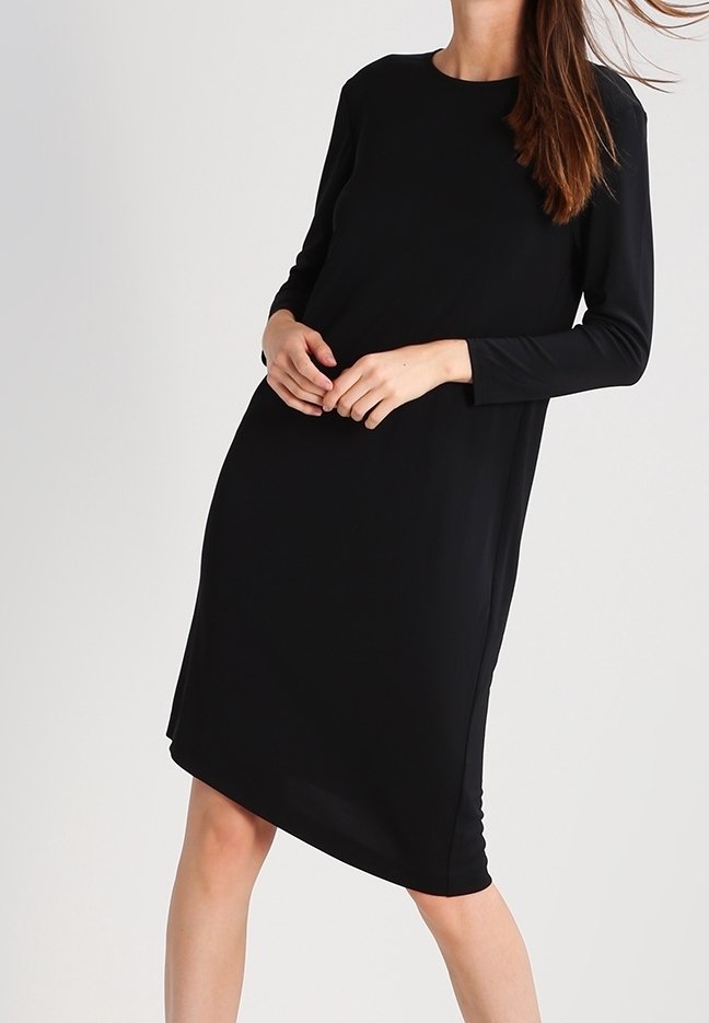 Nainen, jolla on polvimittainen musta pitkähihaisuus mekko, seisoo hieman kääntyneenä ja kädet ristissä edessään yksinkertaista taustaa vasten.