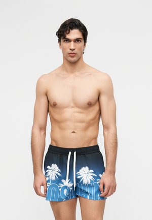 Mladý muž s kučeravými tmavými vlasmi, bez trička, oblečený v modrých plavkových šortkách s bielym potlačou paliem a sťahovacou šnúrkou, stojaci pred jednofarebným pozadím.