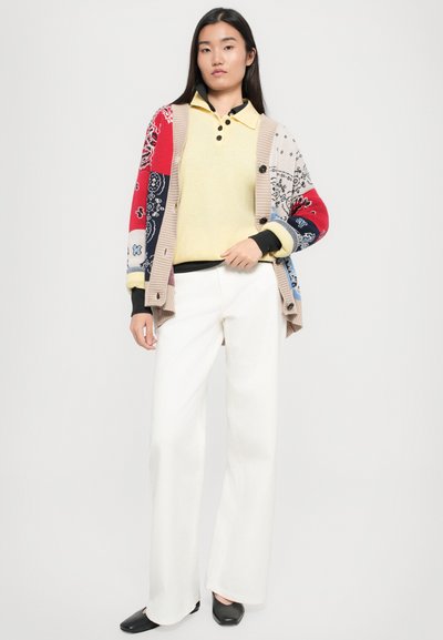 Cardigan multicolore avec des motifs, pull léger jaune, et pantalon large blanc, associé à des chaussures noires à enfiler.