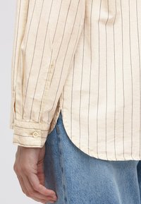 Light beige button-up shirt with dark vertical stripes, featuring cuffed sleeves and side slits, paired with light blue denim.