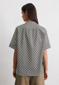 Chemise à manches courtes avec un motif géométrique noir et blanc, col et coupe décontractée. Fabriquée en tissu léger avec une texture lisse.