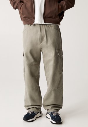 Personne portant un pantalon cargo gris clair, une veste zippée marron, un t-shirt blanc et des baskets noires et blanches, debout devant un fond uni.