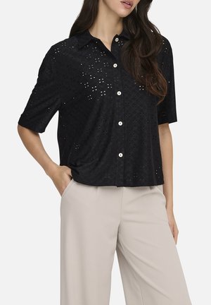 Femme portant une chemise noire à manches courtes boutonnée avec motif à œillets et un pantalon beige taille haute à jambes larges, main dans la poche.