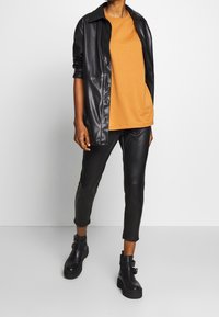 Černá bunda z umělé kůže, oranžové tričko a černé kalhoty z umělé kůže. Robustní černé kotníkové boty s přezkami. Hladké textury, moderní design.