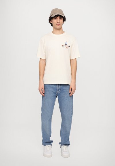 Beige t-skjorte med grafisk trykk, lyseblå jeans og en lys beige caps. Modellen står med hendene på sidene foran en ensfarget hvit bakgrunn.
